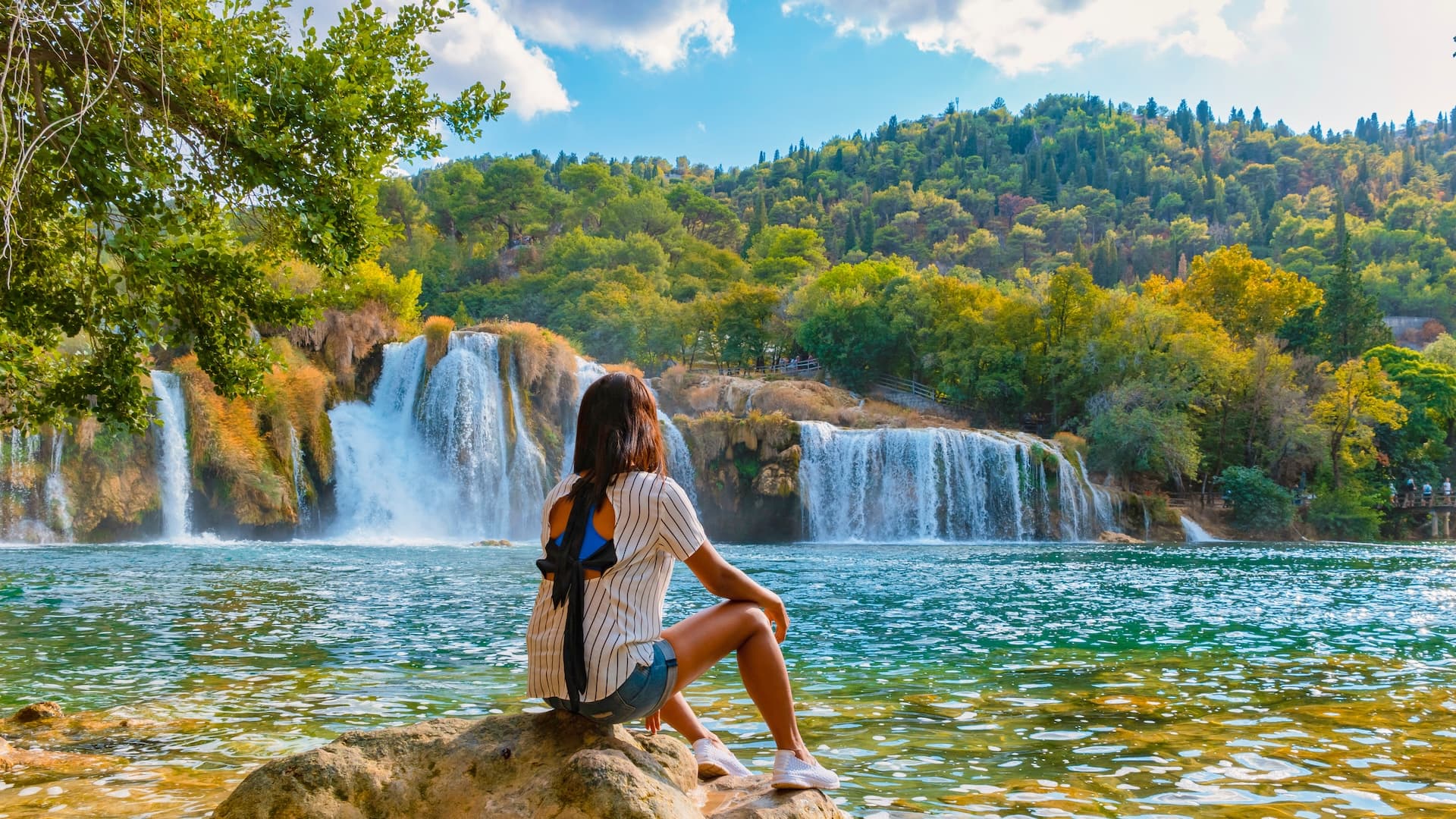 Woman sitting on rock watching Krka waterfalls cascading into turquoise water pool