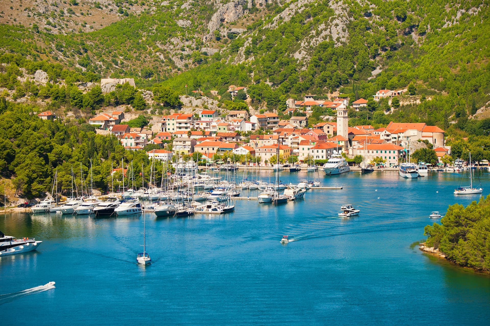 Boats in a harbor next to a coastal town with terracotta roofs backed by green mountains.