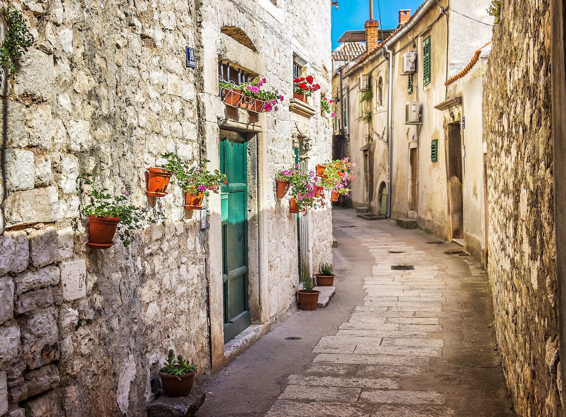 Narrow cobbled street with stone walls, green door, and potted flowers in a sunny historic town.