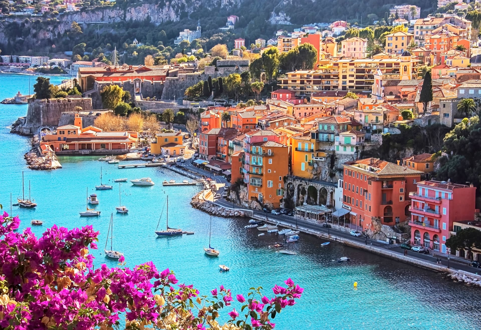 Sailboats moored in turquoise harbor by colorful coastal town and steep hillside.