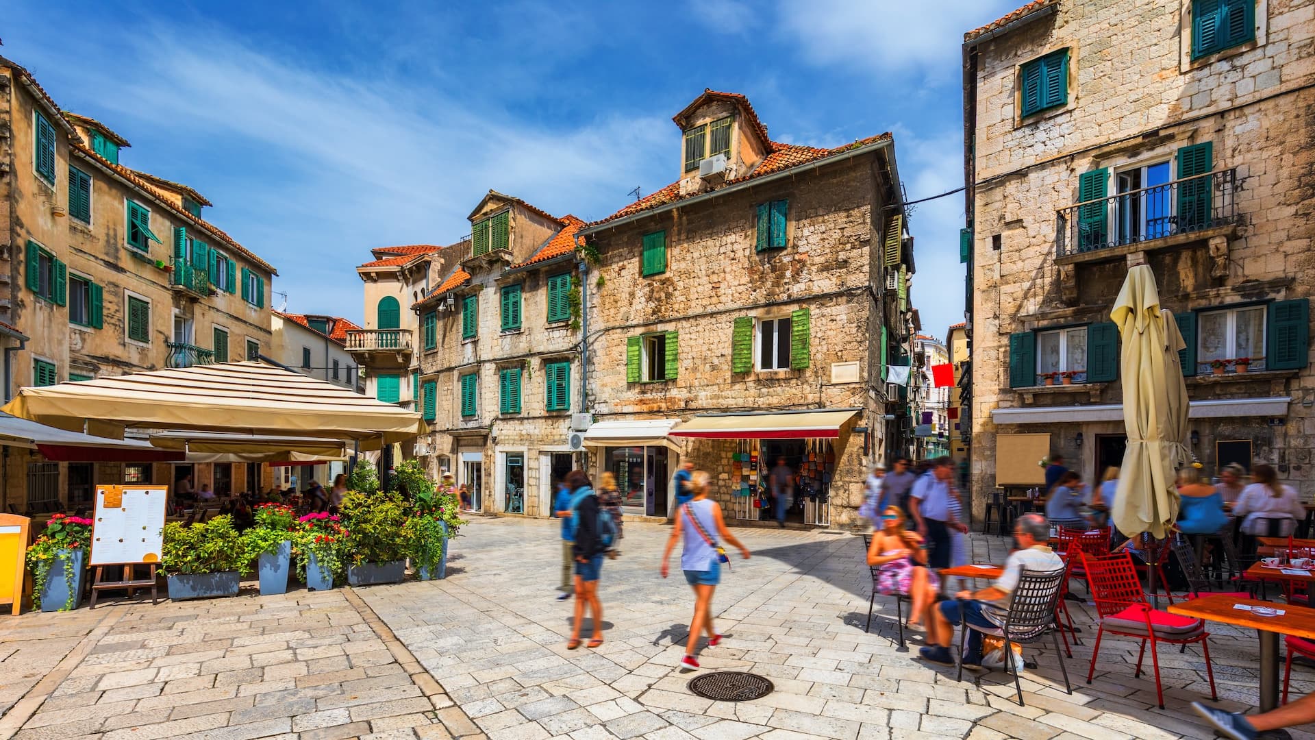 Outdoor cafe dining in a town square with stone buildings and green shutters under a blue sky.
