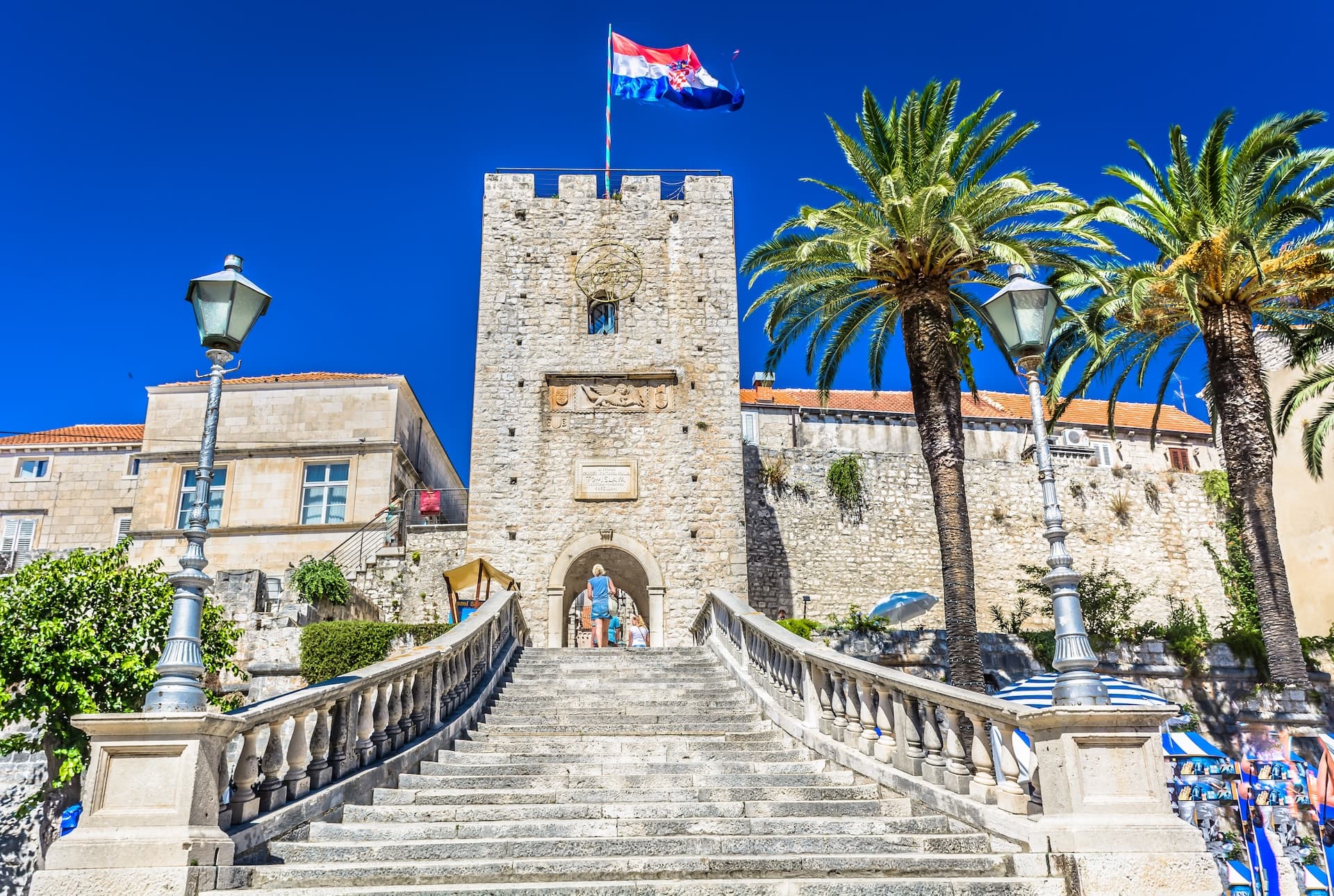 Stone staircase leading to historic tower with Croatian flag, palm trees, Korčula.