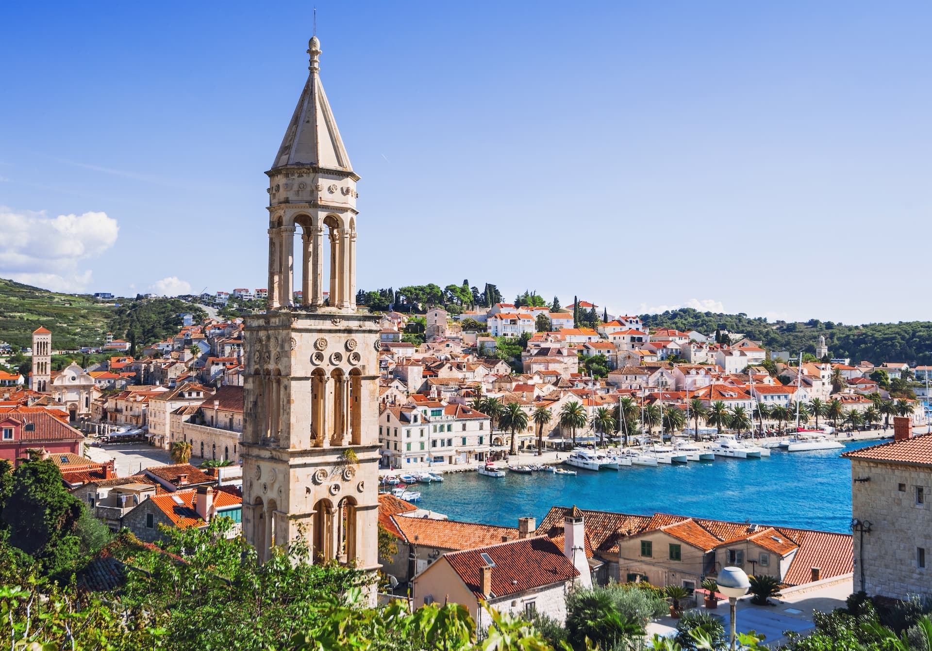 Stone bell tower overlooking Hvar town, red roofs, palm trees, and yachts in the blue harbor.