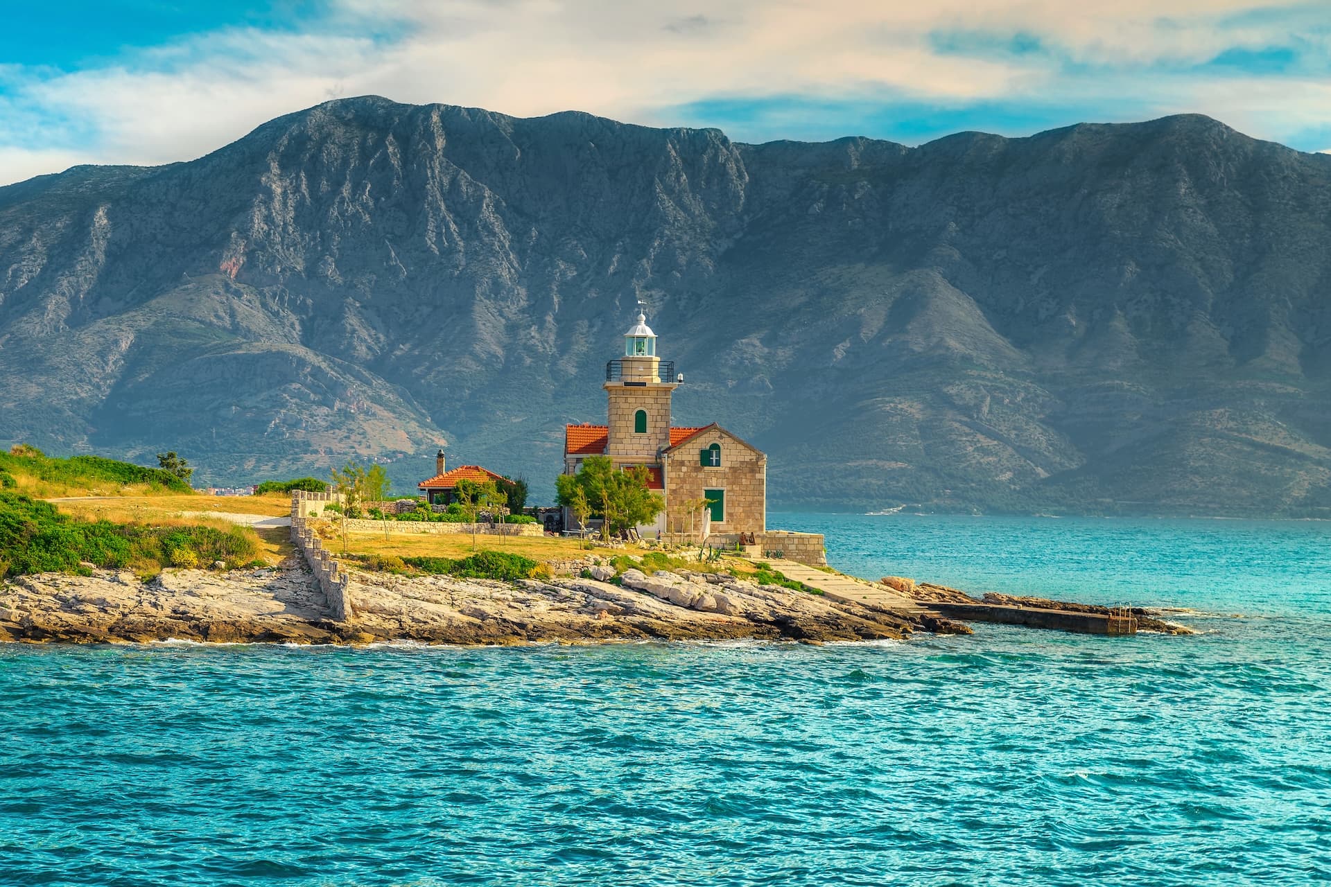 Stone lighthouse on rocky coast with turquoise water and large mountain backdrop, Hvar Island.