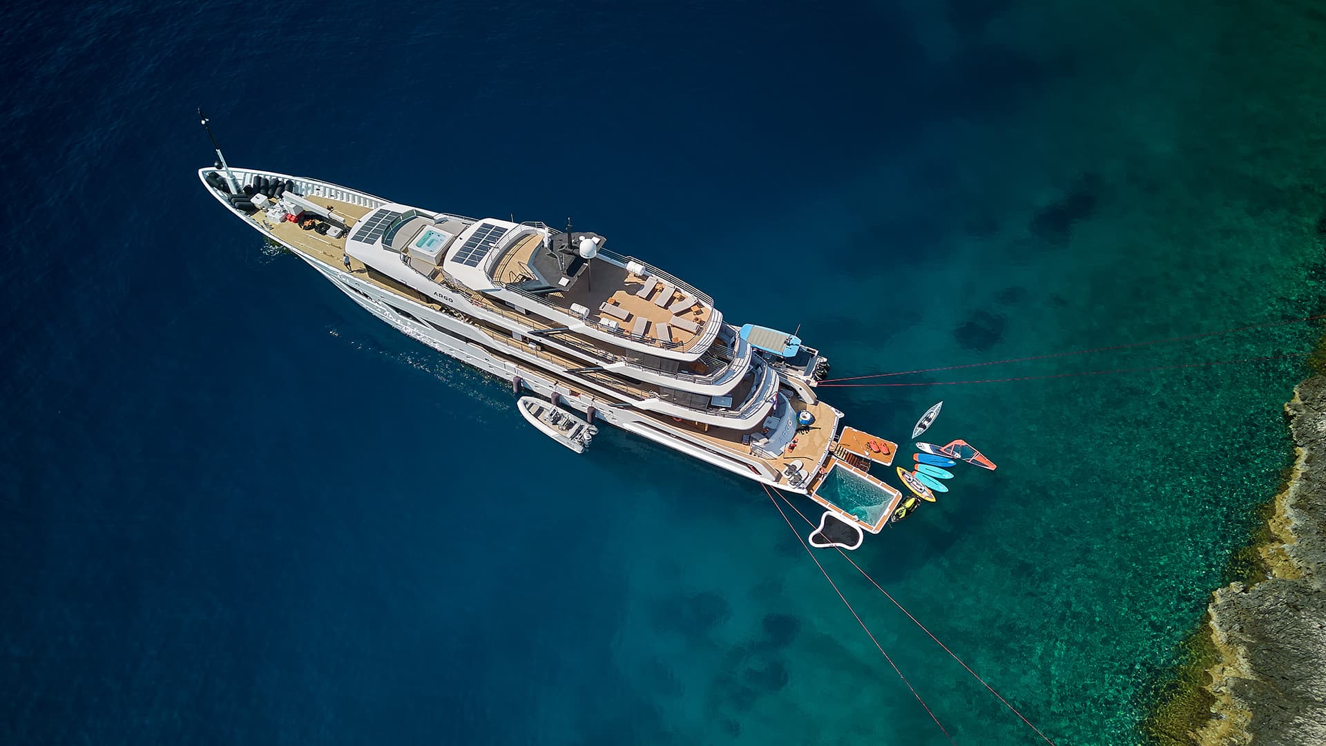Aerial view of large white yacht named ARGO anchored near rocky coastline in clear blue-green water.