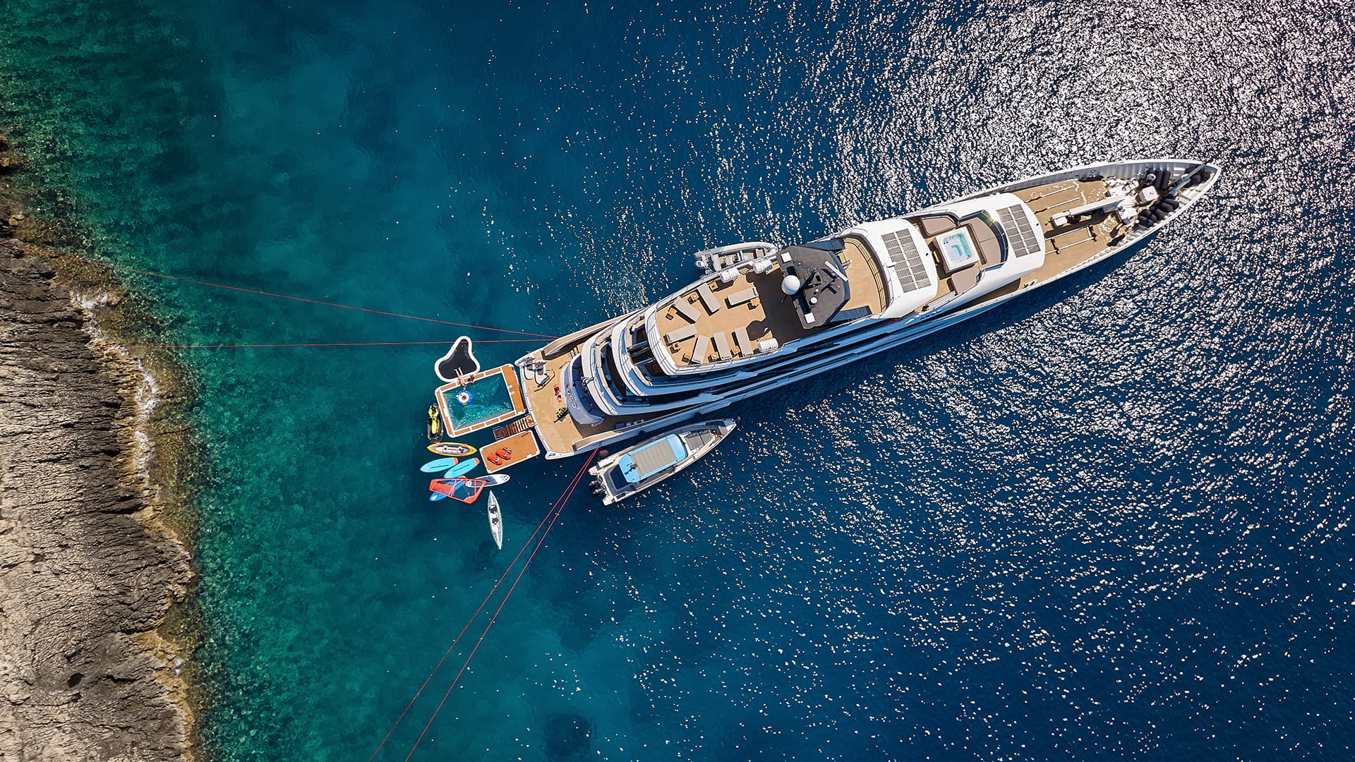 Aerial view of large yacht anchored near rocky coastline with inflatable pool and water toys.