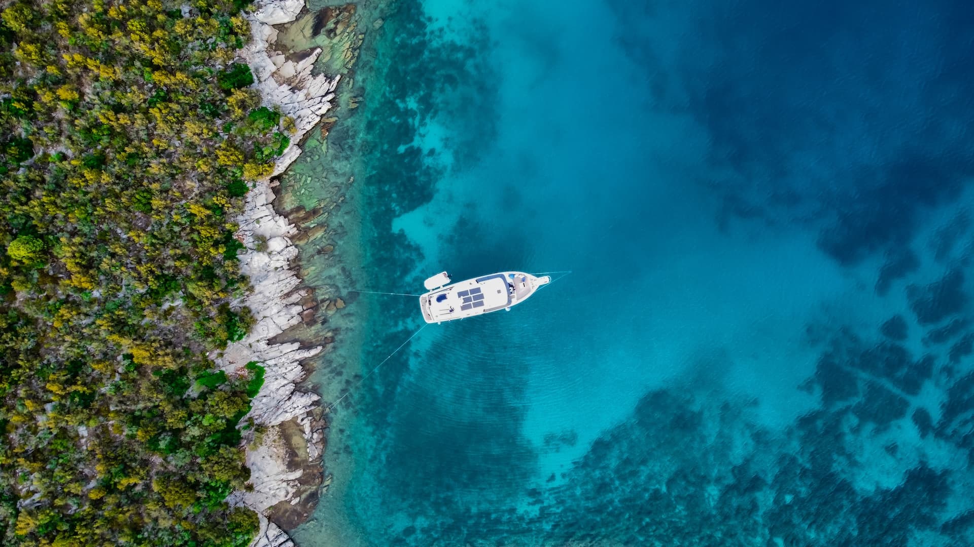 Aerial view of white boat anchored near rocky, green coastline in clear turquoise sea water.