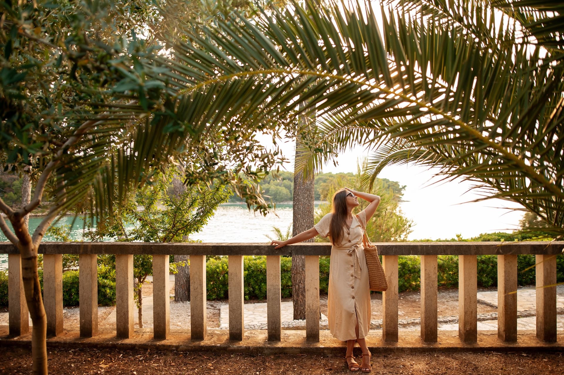 Woman in linen dress by stone railing overlooking water under palm fronds