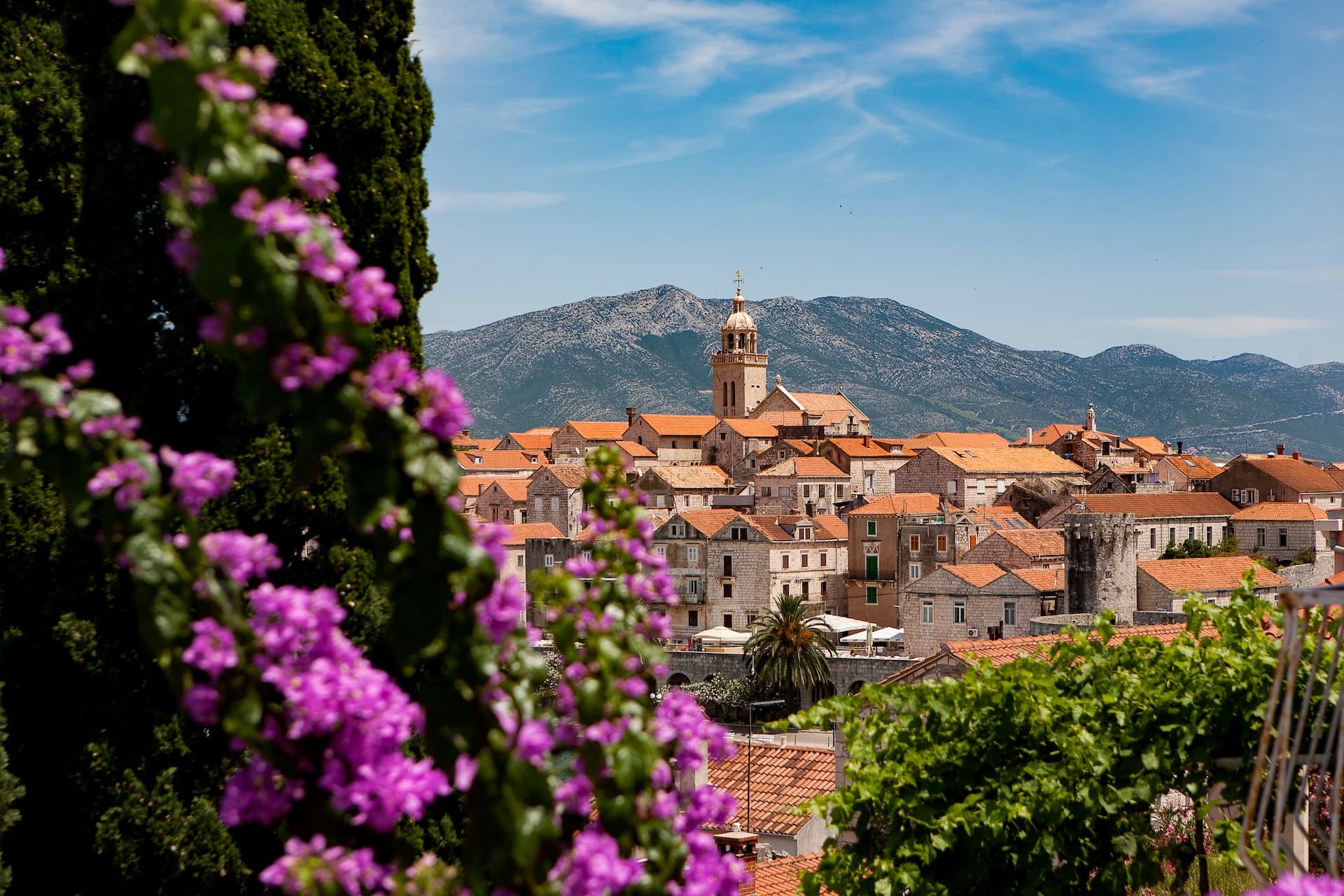 Town with orange roofs and bell tower framed by purple flowers and green foliage in Korčula.