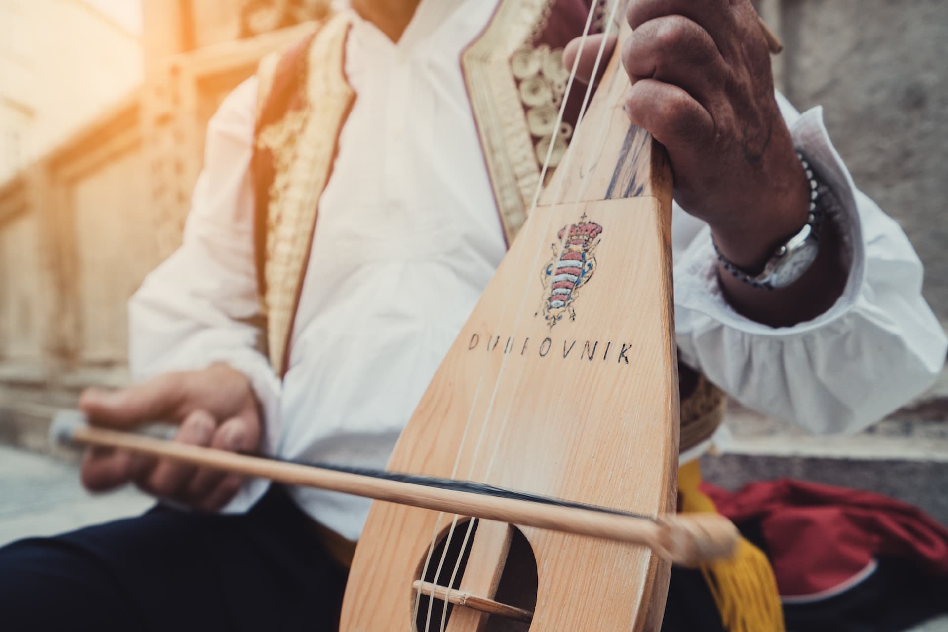 Musician playing a stringed instrument labeled "Dubrovnik" in sunlight.