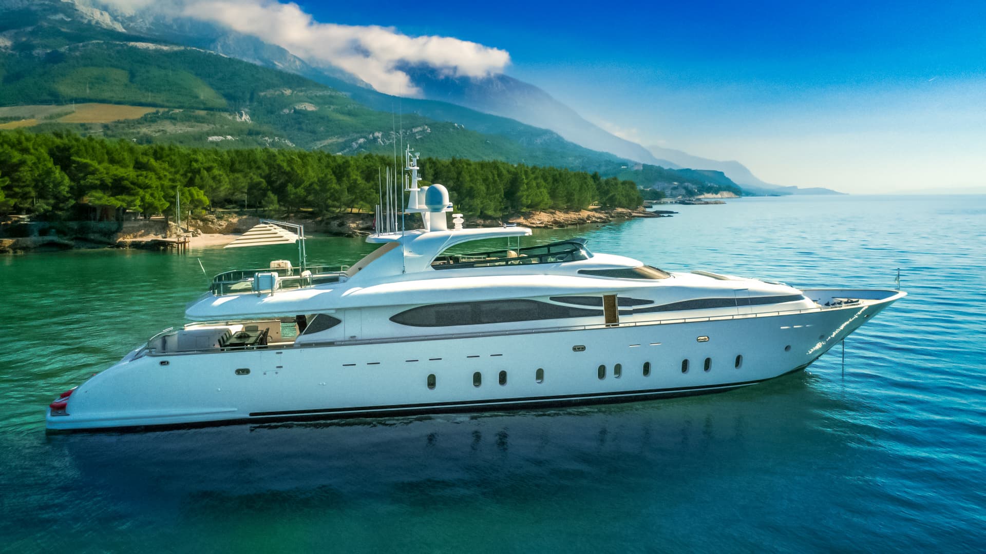 White motor yacht anchored in turquoise water near forested coastline and mountains.
