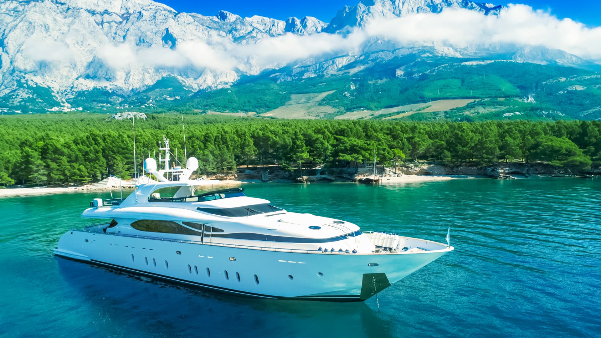 White motor yacht anchored in turquoise water near forested coastline and snow-capped mountains.