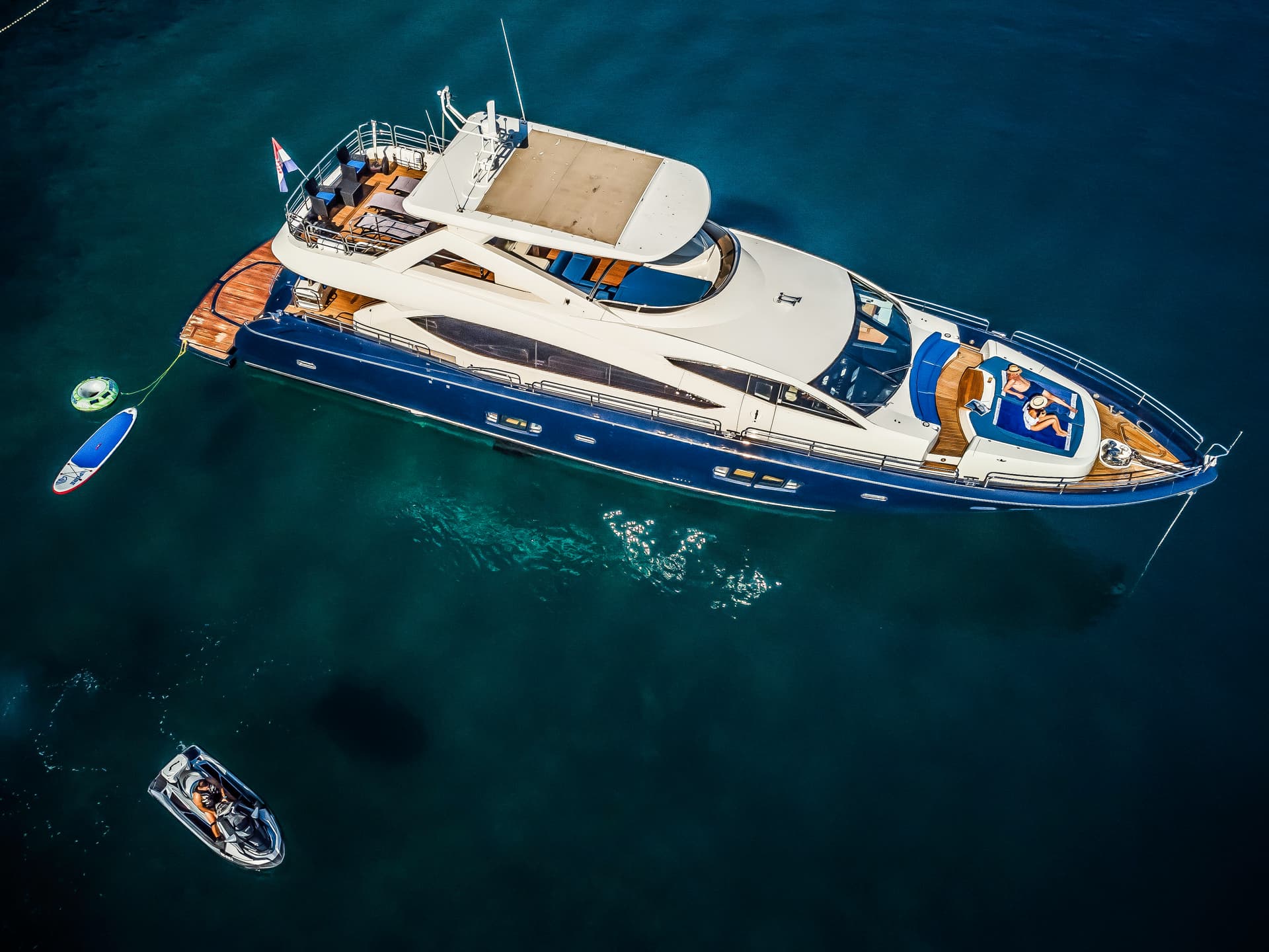 Luxury yacht anchored in dark blue water with paddleboard, inflatable, and jet ski nearby.