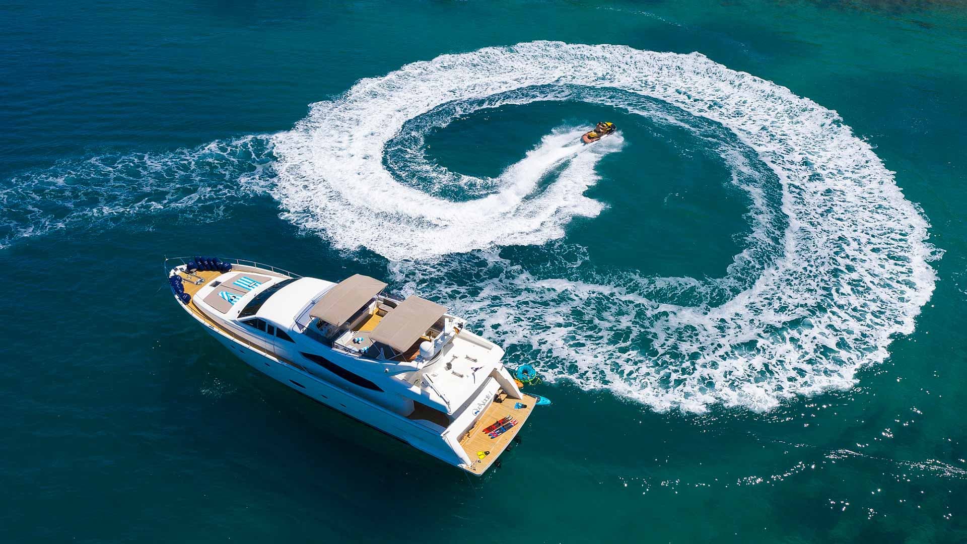Yacht anchored while jet ski makes a circular wake on turquoise coastal water