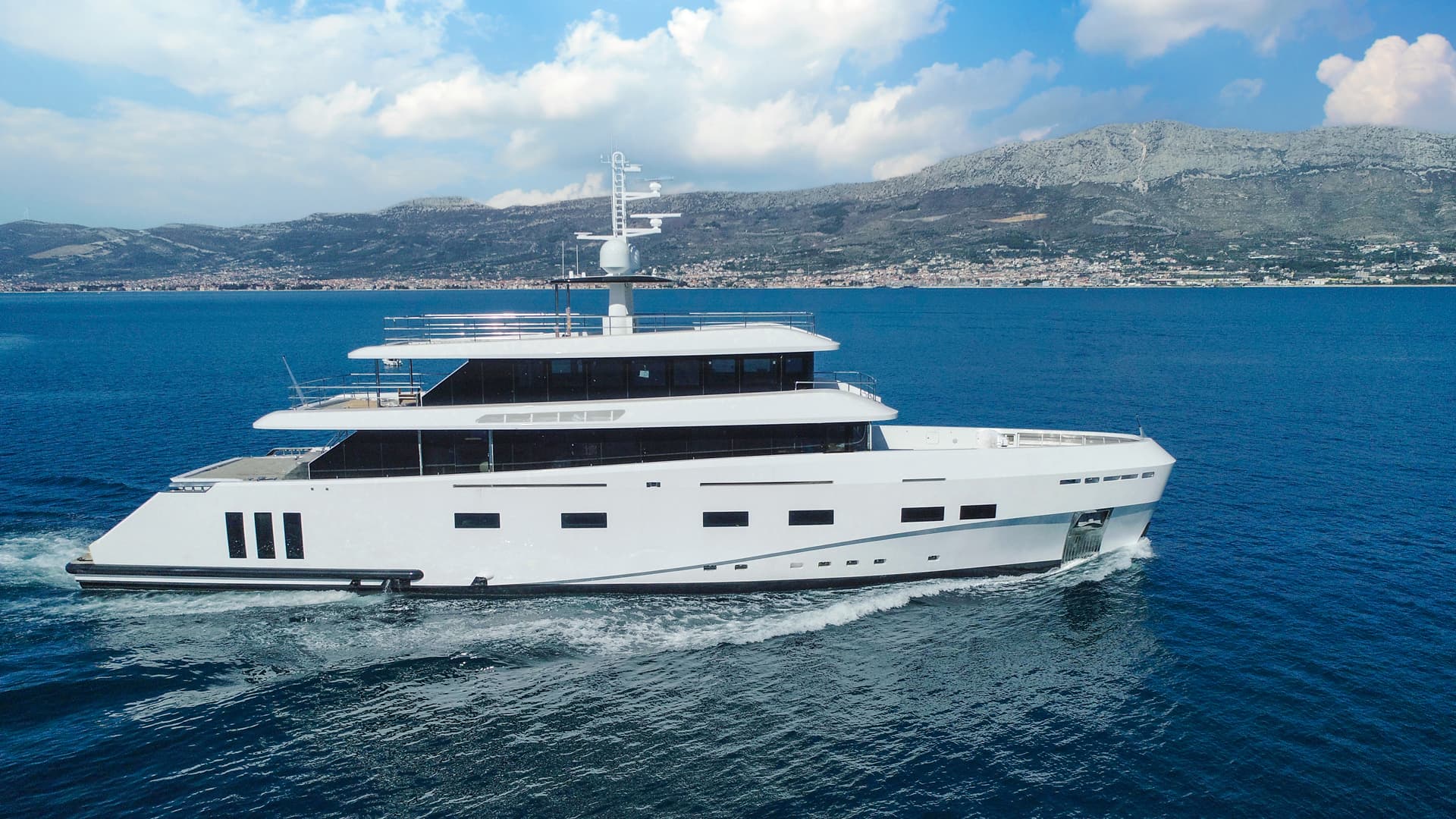 White motor yacht cruising on blue sea with coastal mountains and town in background