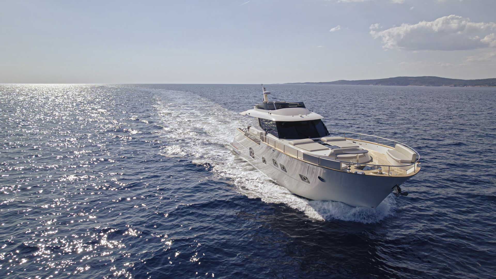Motor yacht speeding through sunlit blue water near a hilly coastline