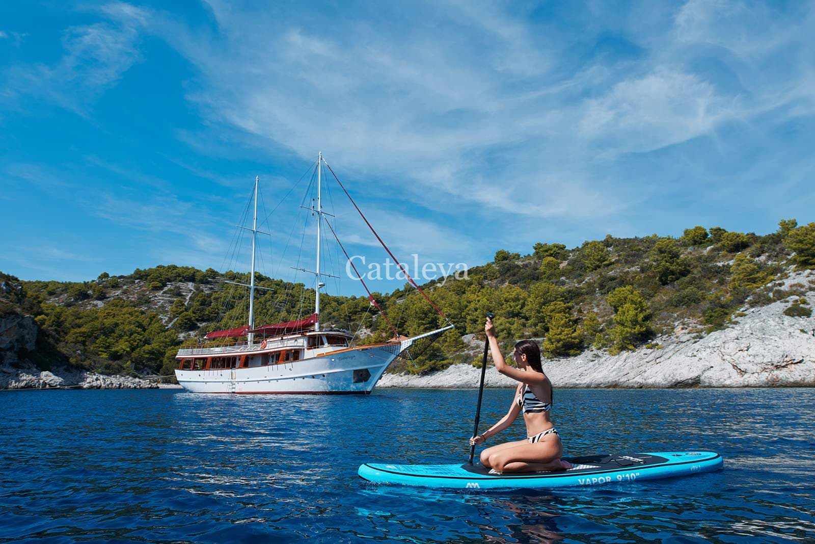 Woman paddleboarding near a large white sailboat off a rocky, green coastline.