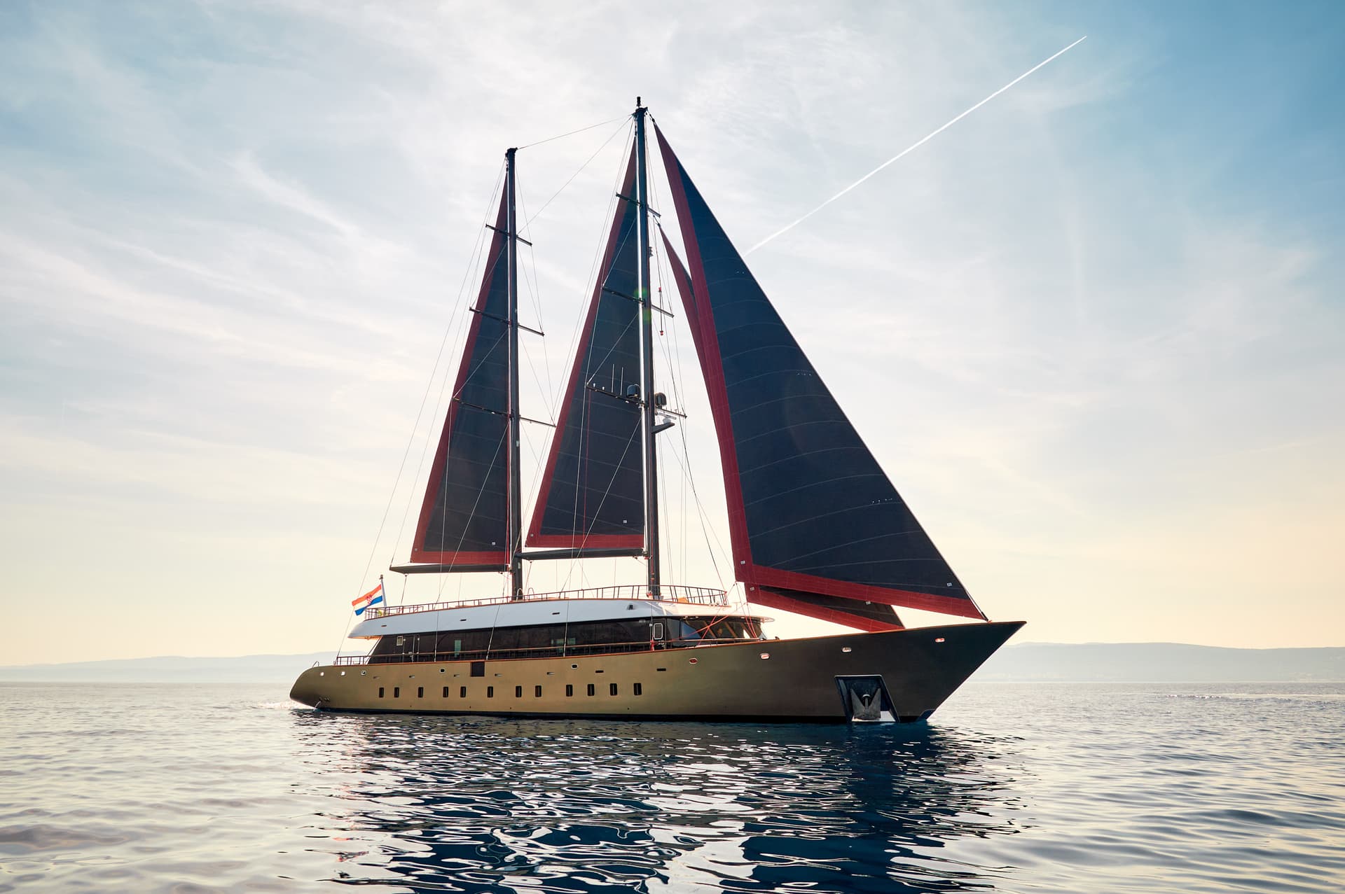 Motor yacht with dark sails on calm water, Croatian flag visible, hazy mountains in background.