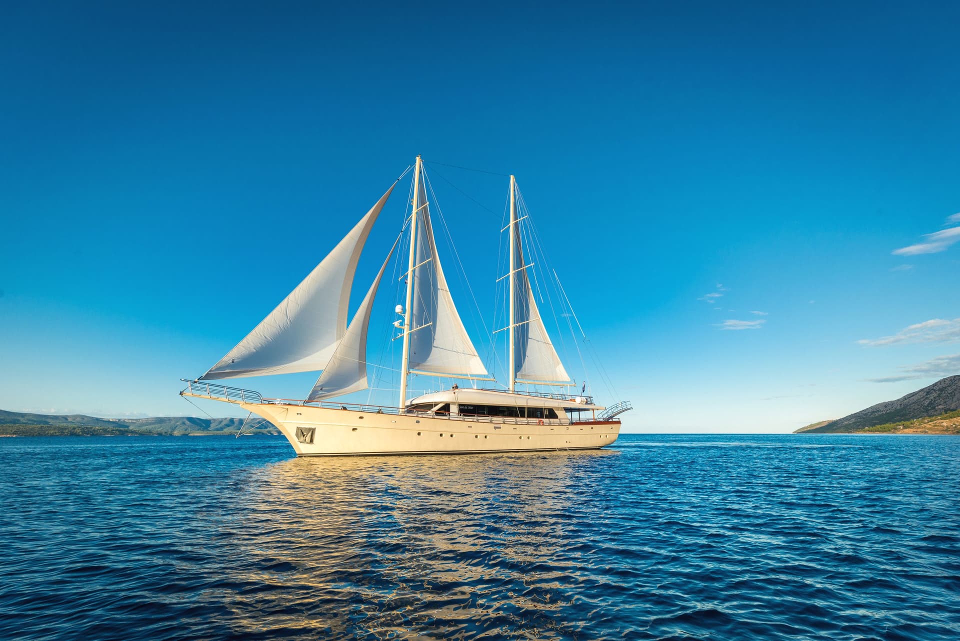 Large sailboat with white sails on deep blue water near a hilly coastline under a clear sky.
