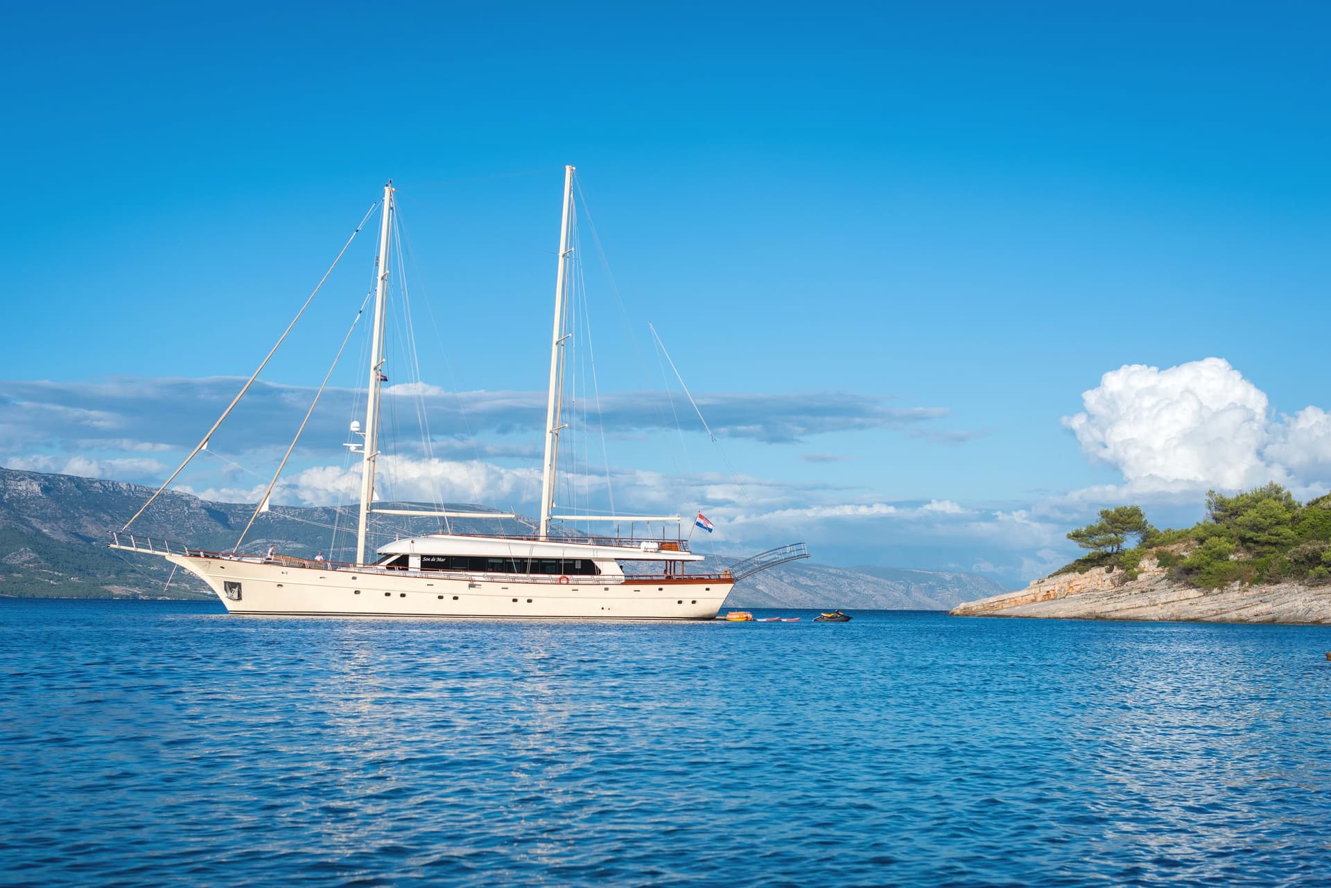 Large sailboat named Son de Mar anchored in blue coastal waters near a rocky, tree-covered shore.
