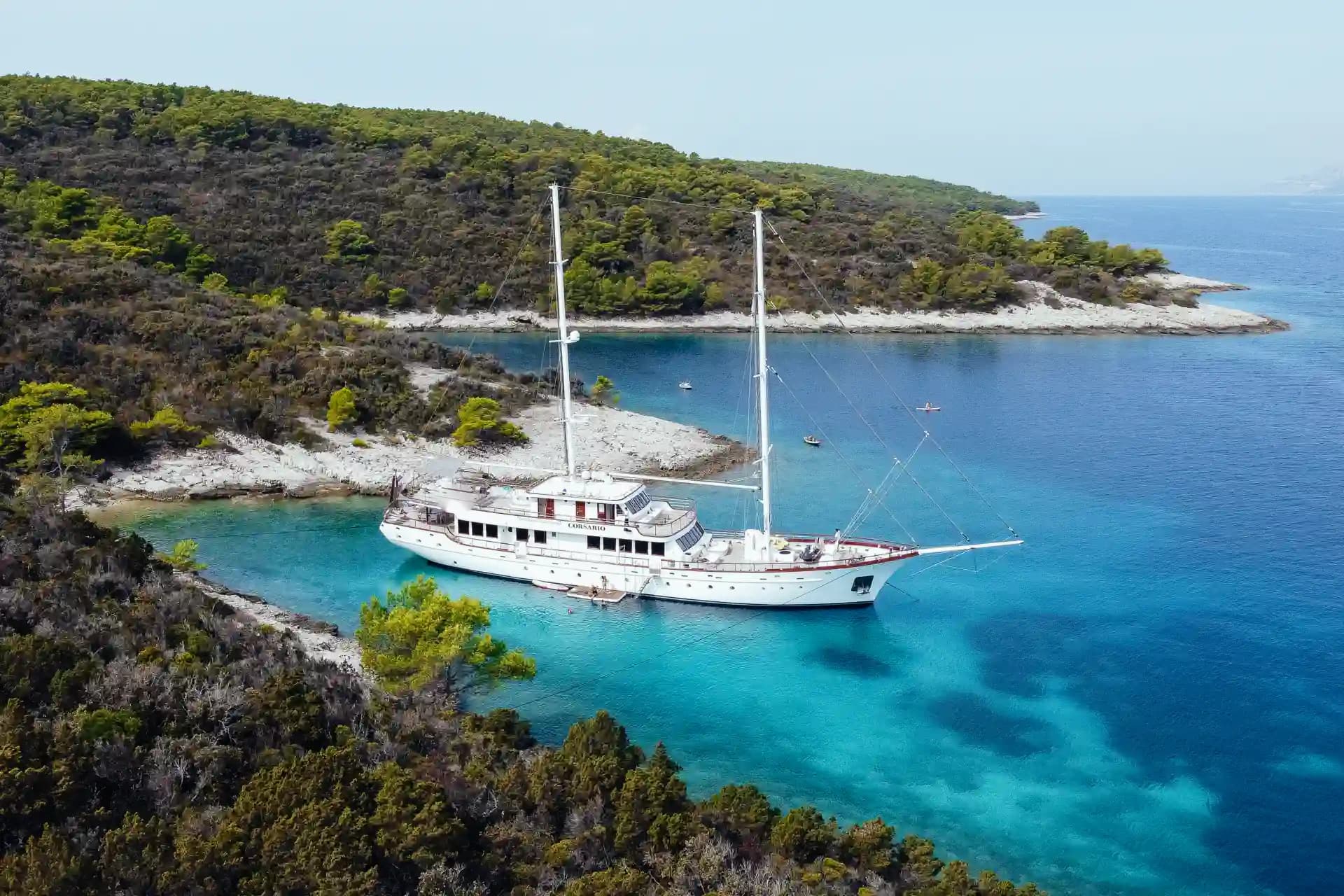 Large white sailboat anchored in turquoise cove near heavily wooded coastline.