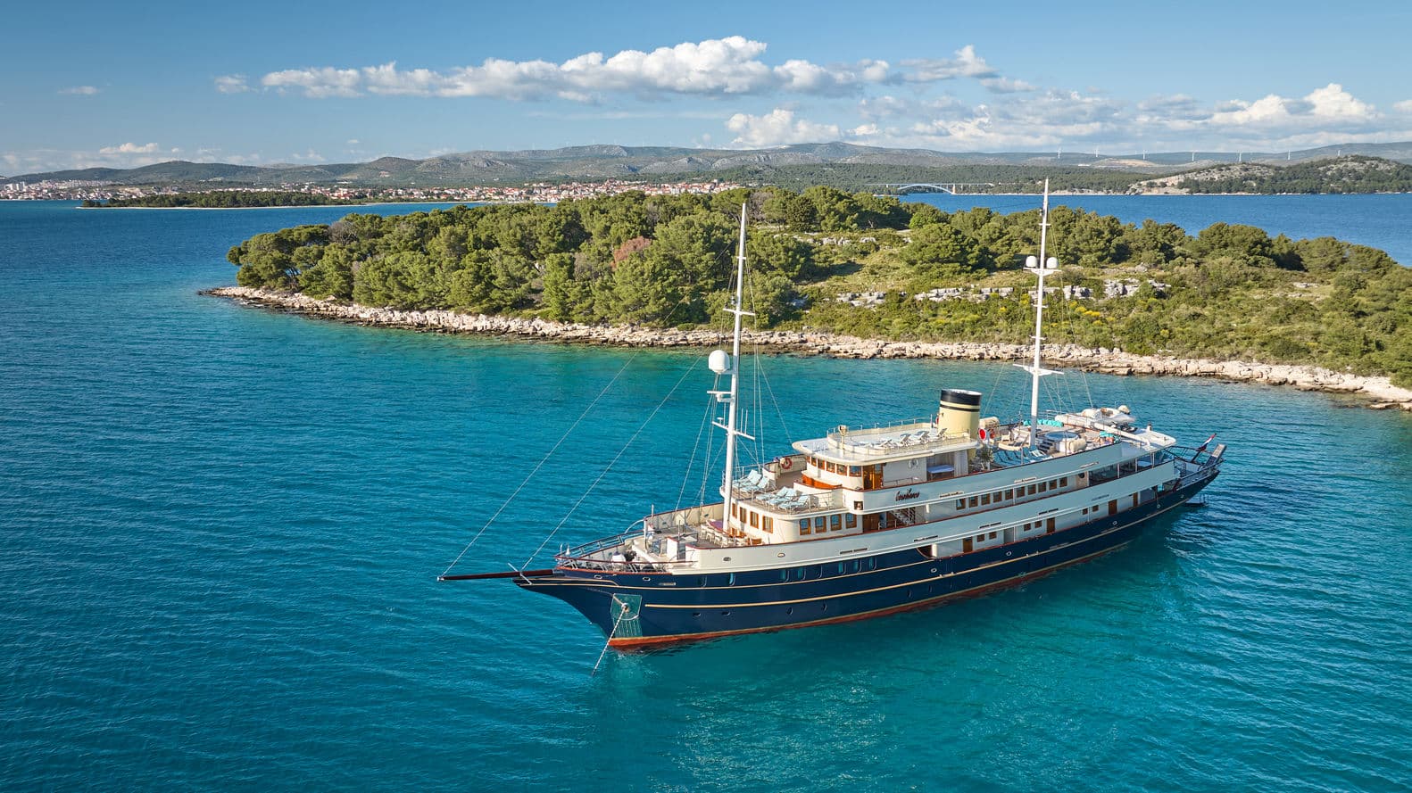 Large yacht anchored near a wooded island with a coastal town and bridge in the background.