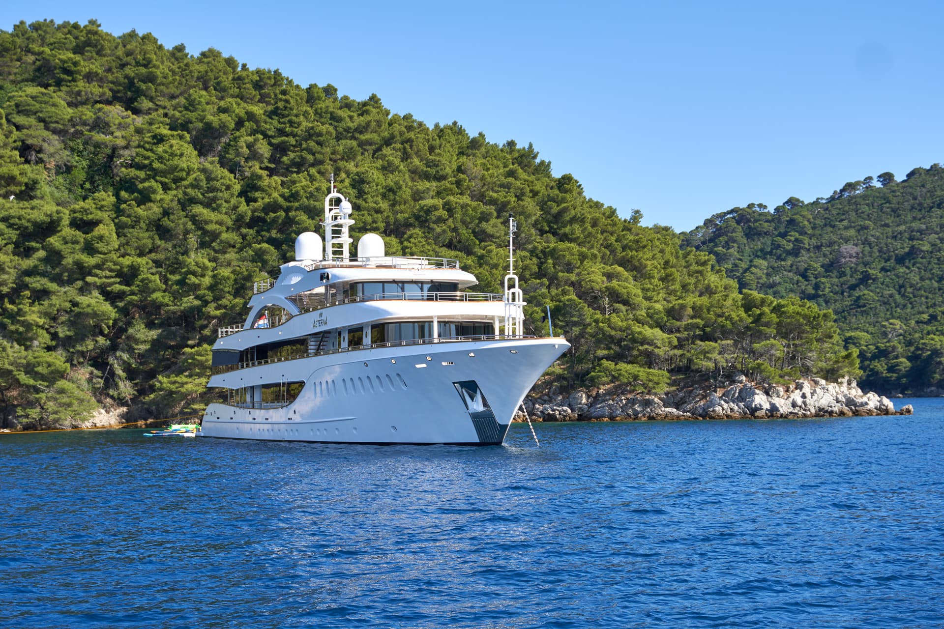 Large white yacht named Aeterna anchored near a densely forested rocky coastline under a clear blue sky.