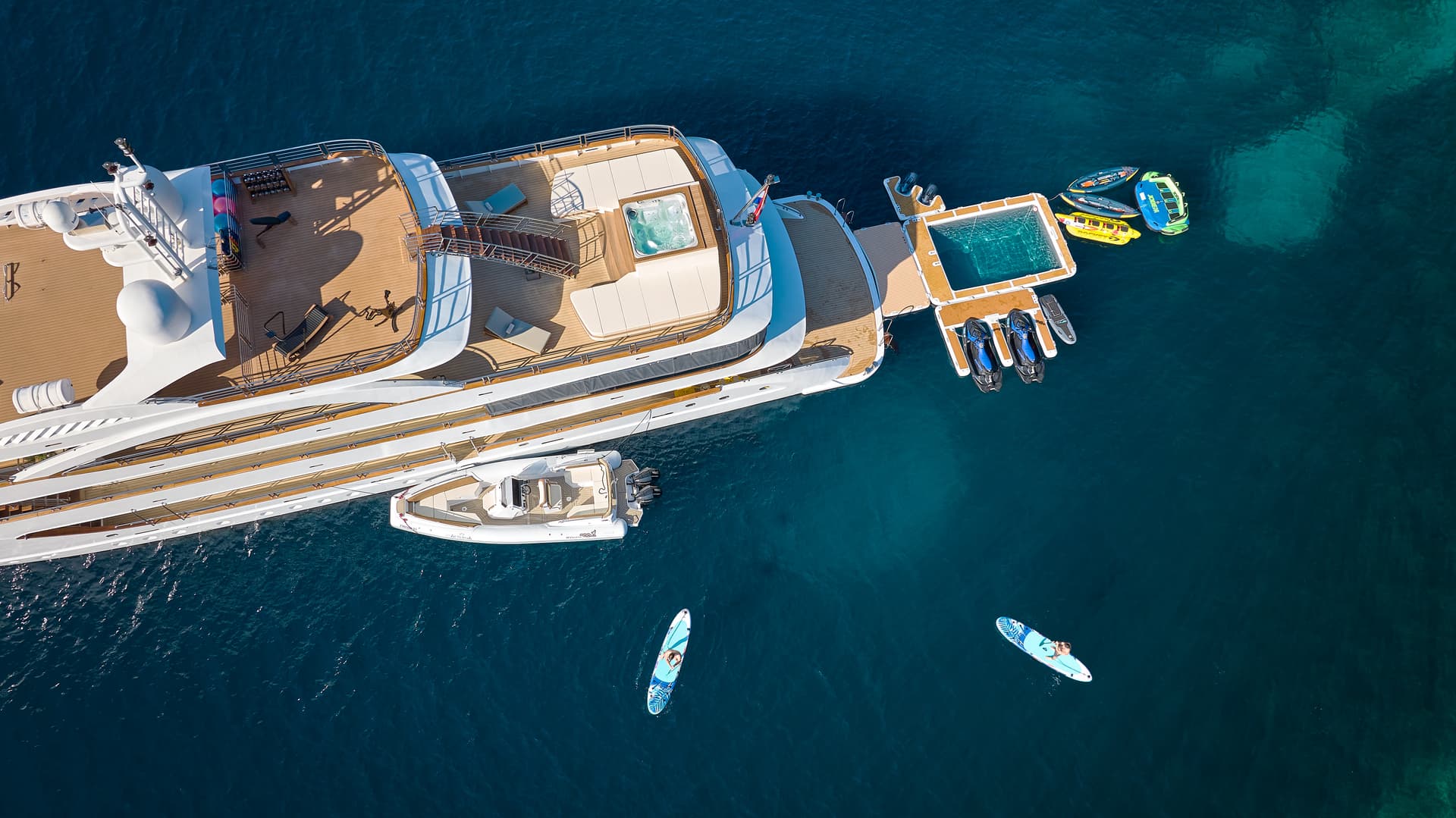 Aerial view of large yacht with jacuzzi, pool, and water toys anchored in deep blue sea.