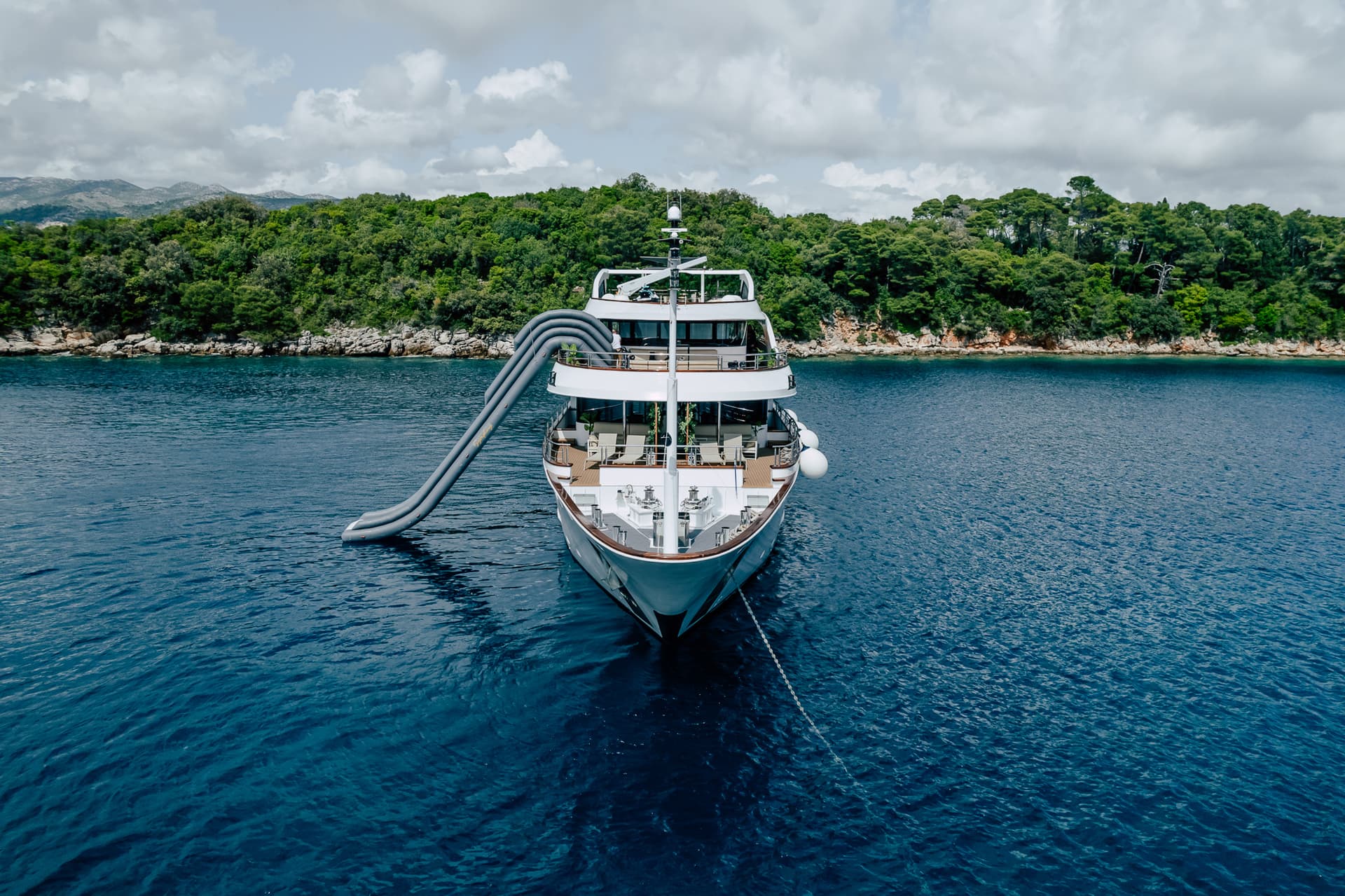 Large white yacht anchored near a lush green coastline with an inflatable water slide deployed.