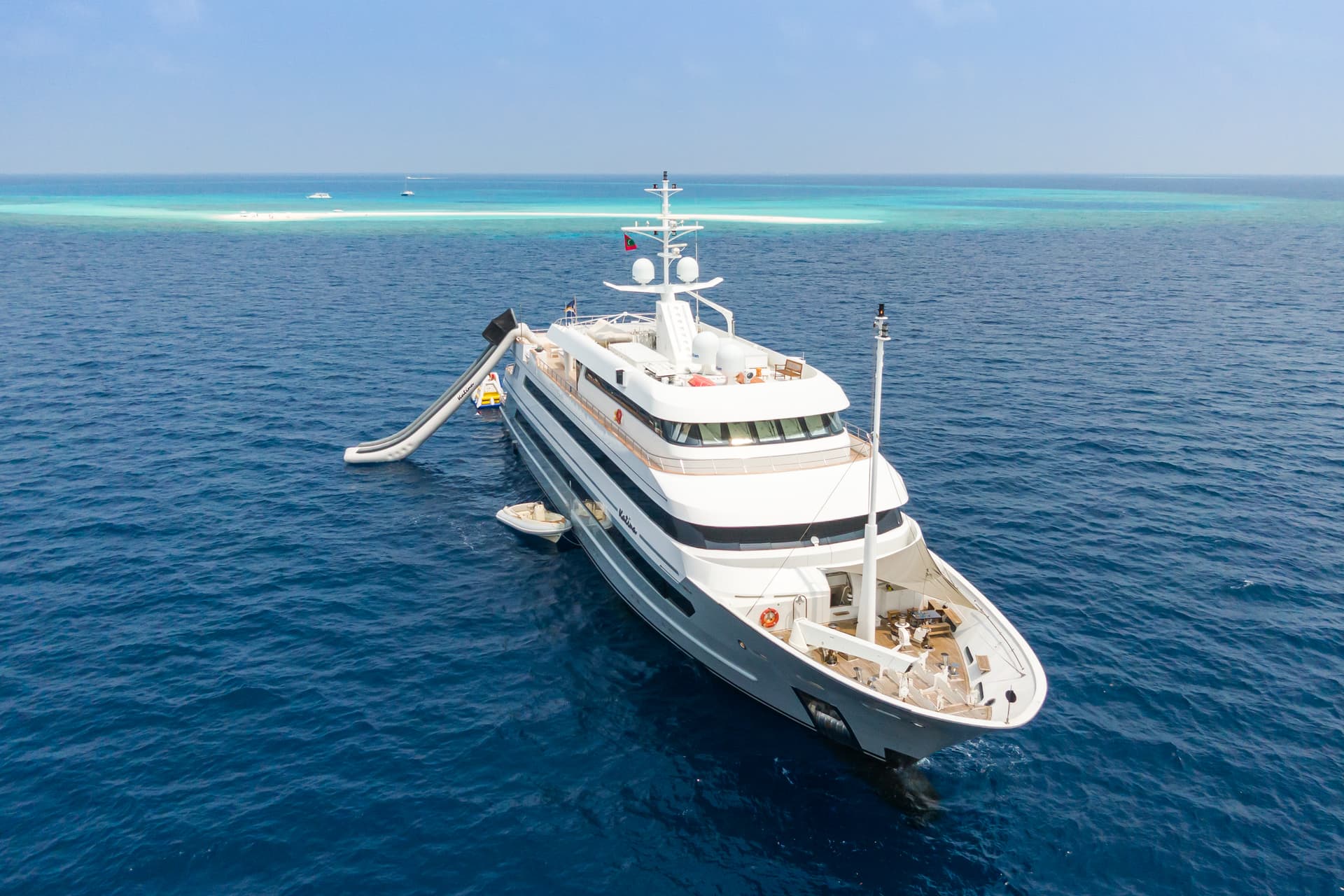 Large white yacht anchored near sandbar with inflatable water slide in deep blue tropical sea.