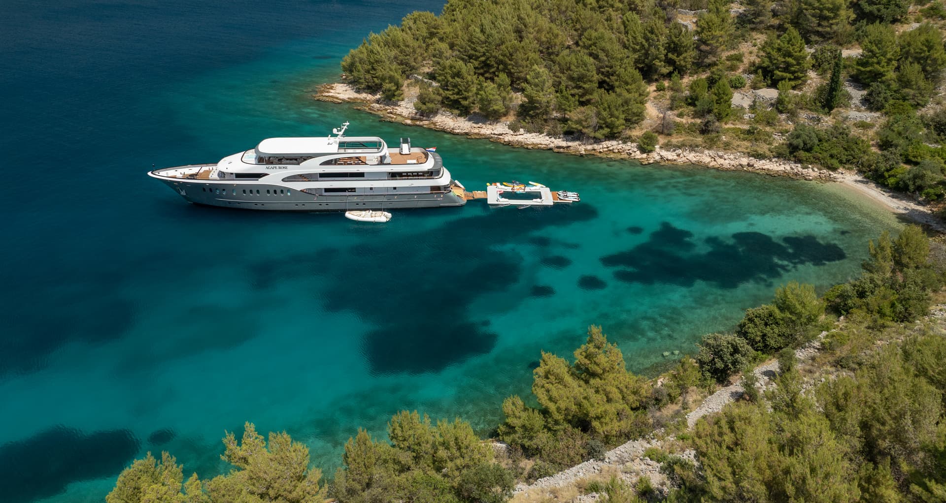 Large yacht anchored in turquoise cove next to rocky, tree-covered coastline.