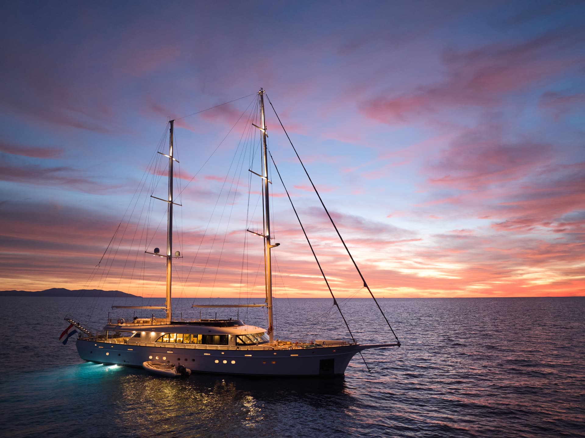 Large sailing yacht illuminated at sunset on dark water with distant mountains and Croatian flag.